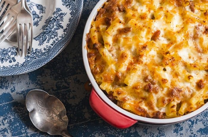 Baked Mac and Cheese With Tomatoes in a medium red casserole dish, serving plates, spoon and forks ready on the side