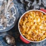 close up of Baked Mac and Cheese With Tomatoes in a medium red casserole dish, serving plates, spoon and forks ready on the side