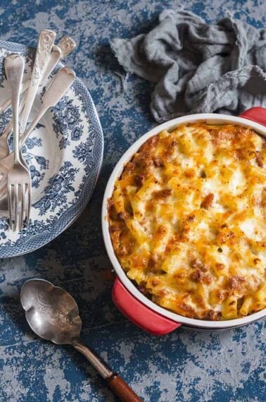 close up of Baked Mac and Cheese With Tomatoes in a medium red casserole dish, serving plates, spoon and forks ready on the side