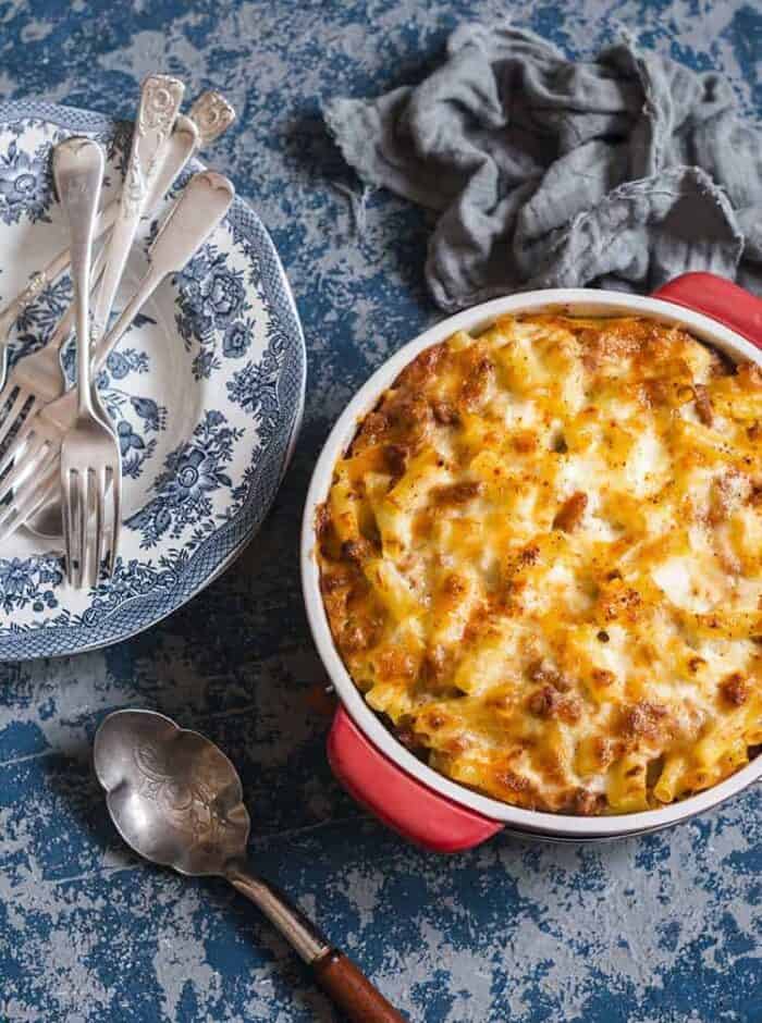 close up of Baked Mac and Cheese With Tomatoes in a medium red casserole dish, serving plates, spoon and forks ready on the side