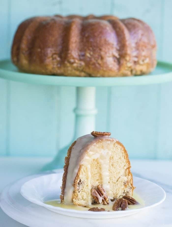 a slice of glazed baked Rum Cake in white dessert plate, whole cake in a cake stand on background