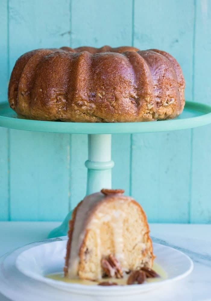 a slice of glazed baked Rum Cake in white dessert plate, whole cake in a cake stand on background