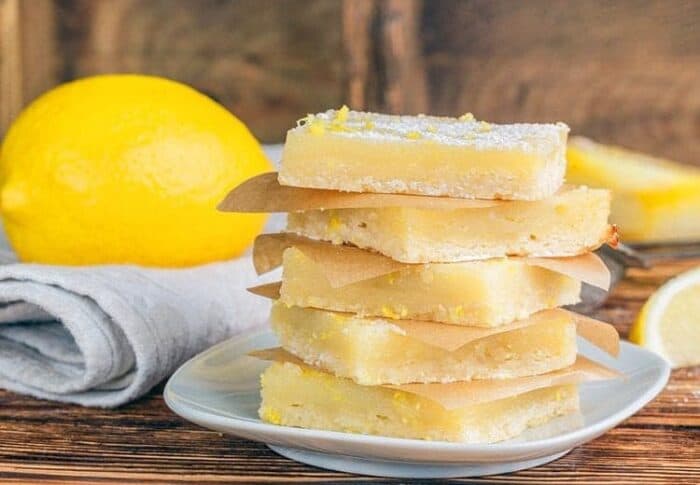 a stack of Creamy Lemon Bar slices in a white dessert plate, fresh lemon over white kitchen cloth on background