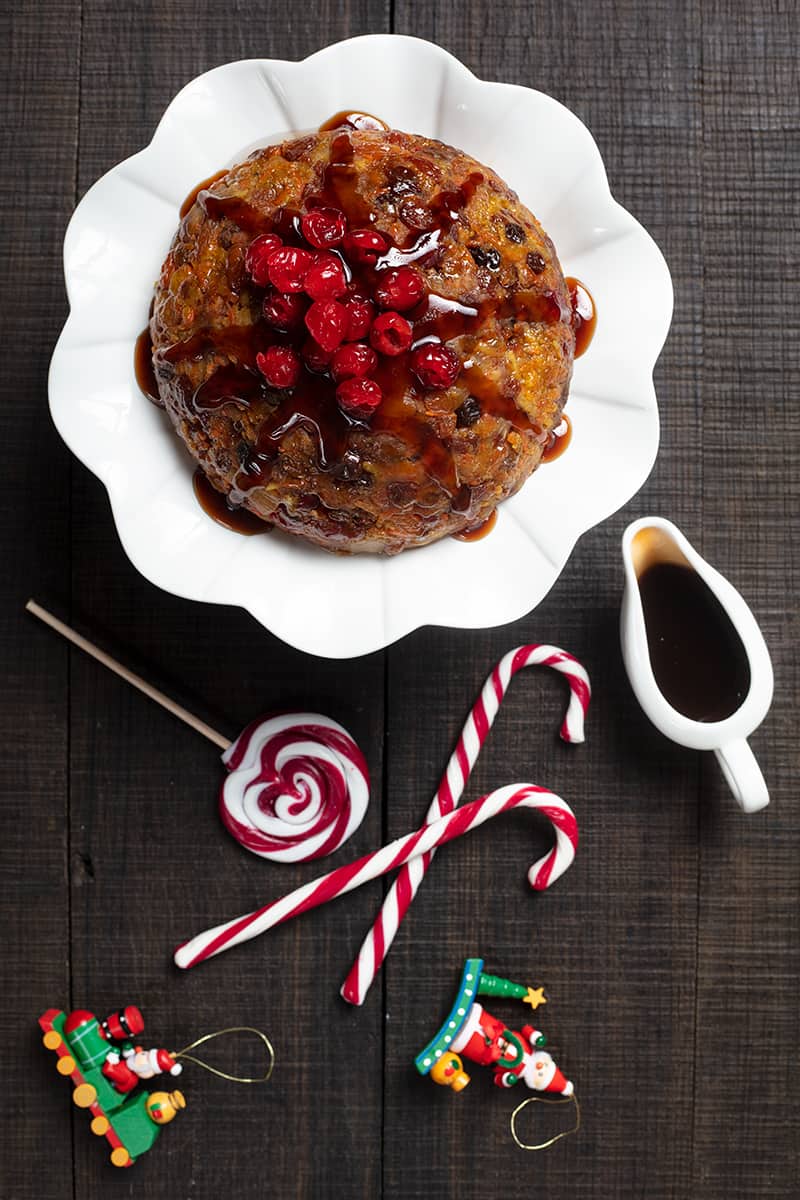 high angle shot of carrot Christmas Pudding on a white ceramic cake holder, brown sugar sauce on white mini pitcher and Christmas candy bars on sides