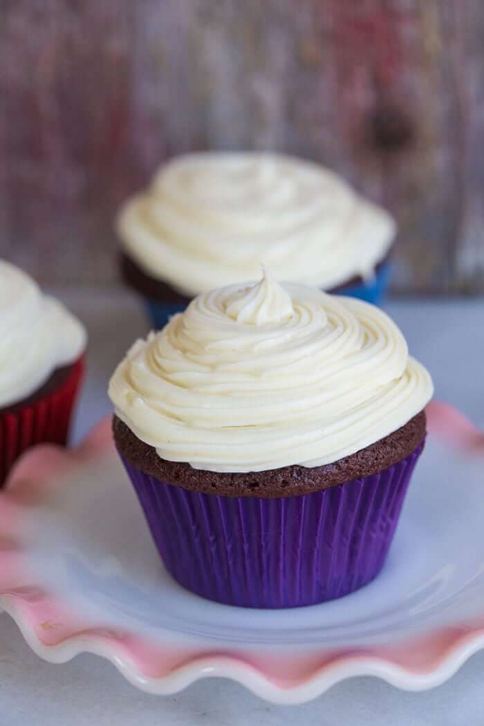 cupcake in a white plate with cupcake liner and Cream Cheese Icing swirled on top, more cupcakes with cream cheese icing on the background