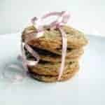 A stack of five chocolate chip cookies is tied together with a curled pink ribbon, resting on a plain white plate against a light background.
