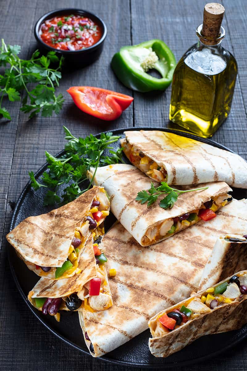 stack of Quesadillas in a large black serving plate garnish with parsley leaves, sliced green and red peppers, oil in bottle and dipping sauce on its background