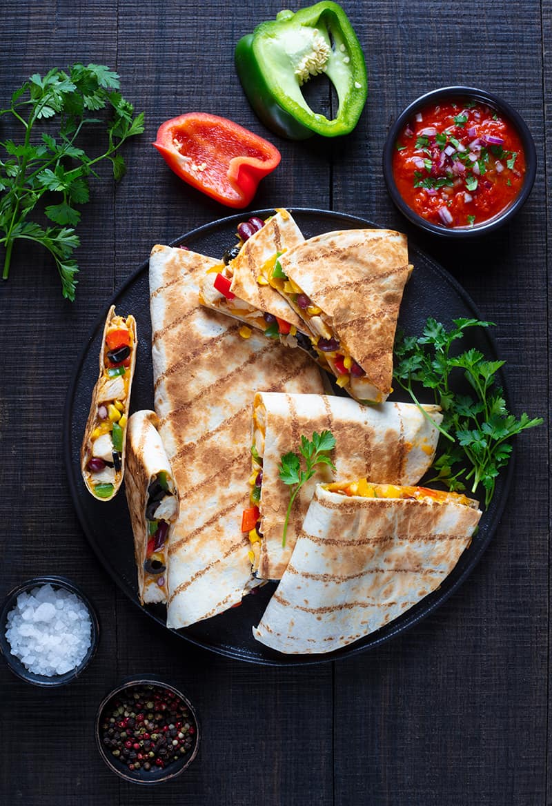 stack of Quesadillas in a large black serving plate garnish with parsley leaves, dipping sauce, slice of red and green peppers beside the plate