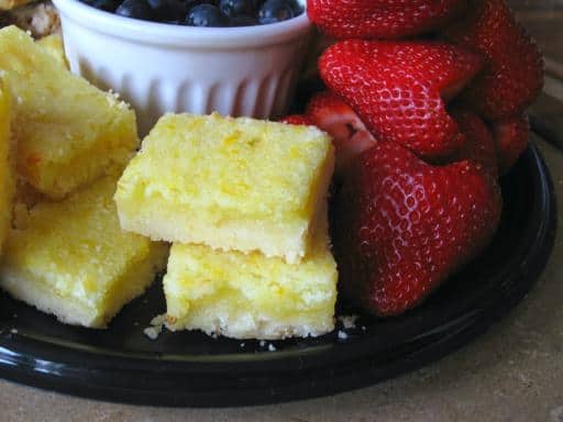 A plate with stacked lemon bars, fresh whole strawberries, and a bowl of blueberries in the background.