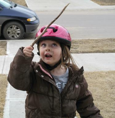 little girl wearing pink bike helmet and brown winter jacket holding a stick
