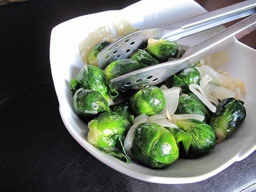 Brussel sprouts in a white bowl with kitchen tongs