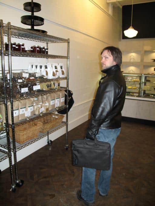 man wearing black leather jacket holding a laptop bag in front of pastry displays