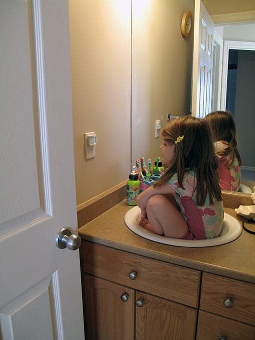 little girl sitting in the sink of the bathroom with a big mirror on her side