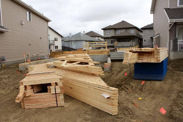 Stacks of wooden boards and construction materials are arranged on a dirt lot between two houses, indicating the early stages of building a new home in a suburban neighborhood.