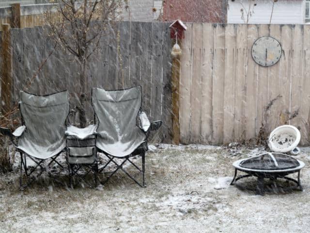 Two folding chairs and a small table sit in a backyard as snow falls. A fire pit with chairs piled on it and a clock on a wooden fence are visible. The ground has a light dusting of snow.