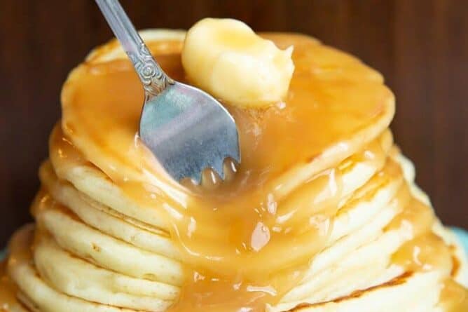 a fork in a stack of Buttermilk Pancakes topped with butter and syrup in a ruffled dessert plate