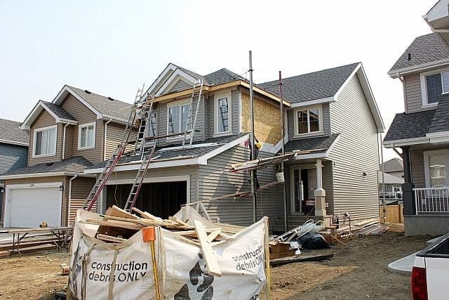 A two-story house under construction with scaffolding and ladders on the front, unfinished exterior siding, and a large bin of construction debris in the yard. Neighboring houses are visible on both sides.