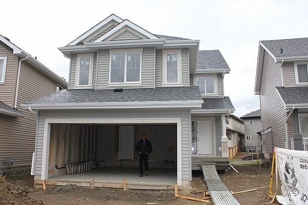A two-story, light gray house under construction with an open garage and unfinished driveway. A person stands inside the garage. Nearby houses and construction materials are visible.