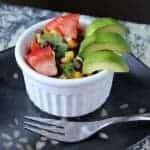 A small white ramekin filled with a vibrant Corn Salad of tomatoes, avocado slices, corn, black beans, and cilantro, placed on a black plate with a silver fork beside it.