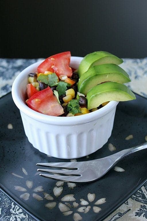 close up of Avocado, Black Bean & Corn Salad in white ramekins on a black plate with fork on side