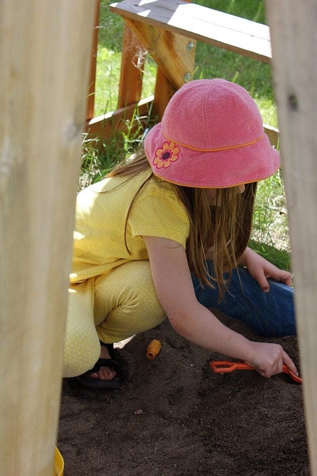 little girl wearing yellow clothes and pink cap playing in the sandbox near the fence