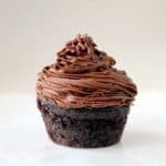 A single chocolate cupcake with rich, swirled chocolate buttercream frosting sits on a plain white surface against a light background.