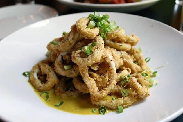 A white plate filled with crispy fried calamari rings, garnished with chopped green onions and served with a light yellow sauce.
