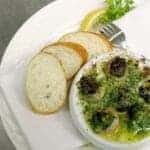 close up of Escargot Forestiere with garlic and melted butter, slices of French bread on side garnish with a slice of lemon at parsley