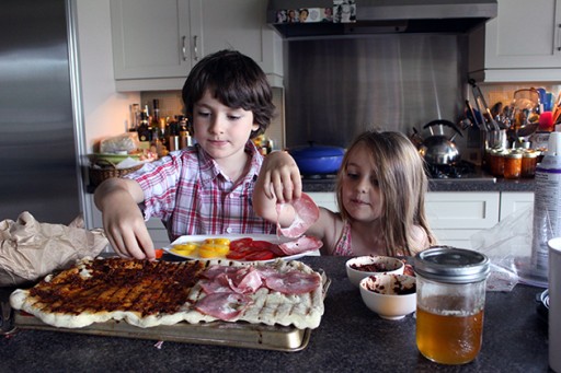 two kids adding the pizza sauce and toppings to the big dough