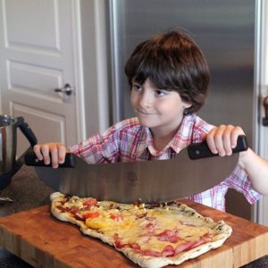 young boy holding a large semi circular stainless pizza knife over the cooked pizza