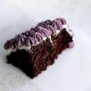 A slice of gluten free chocolate cake with white frosting and decorative purple icing swirls on top, placed on a white plate.