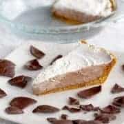 A slice of creamy chocolate mousse pie with a whipped topping sits on a white plate surrounded by broken pieces of chocolate, with the rest of the pie visible in a glass dish in the background.