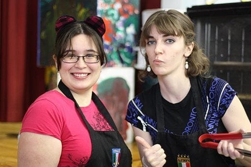 close up shot of two women wearing pink and blue with one holding a red kitchen tongs