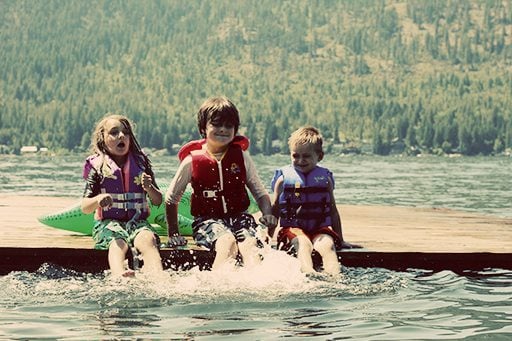 three kids wearing life jackets, sitting down on a dock starting to splash water