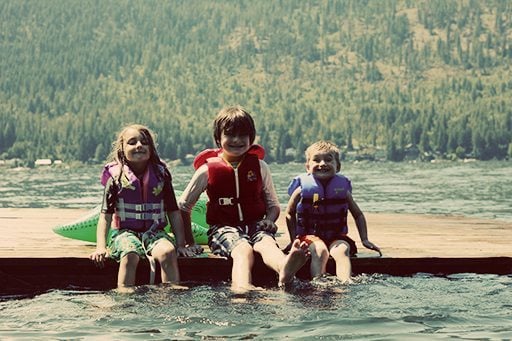 three kids wearing life jackets, sitting down on a dock