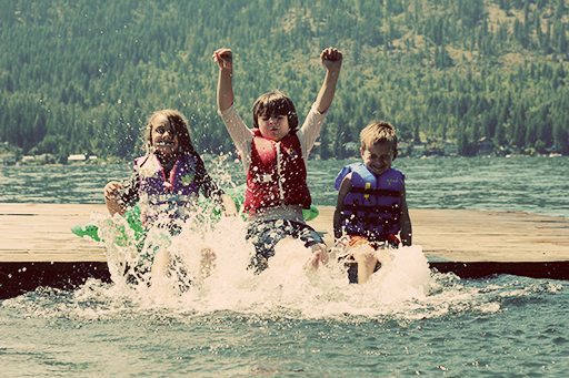 three kids wearing life jackets, sitting down on a dock splashing water using their feet