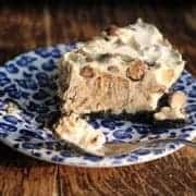 A slice of creamy Ice Cream Peanut Butter Pie with chocolate pieces sits on a blue and white patterned plate, with a fork beside it on a wooden table.