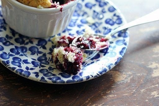 close up spoon of rich Saskatoon berry filling with the cakey biscuit topping on blue saucer plate beside white ramekins