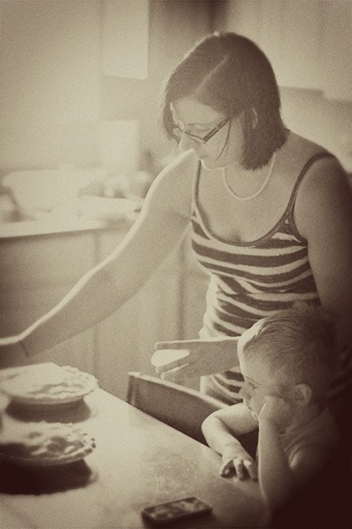 A woman in a striped tank top stands beside a young child at a table, preparing Saskatoon pie. The image has a soft, sepia-toned, vintage effect, with light spilling into the cozy kitchen setting.