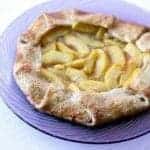 A rustic peach galette with a golden, flaky crust and juicy peach slices arranged in the center, served on a purple glass plate for a charming touch.