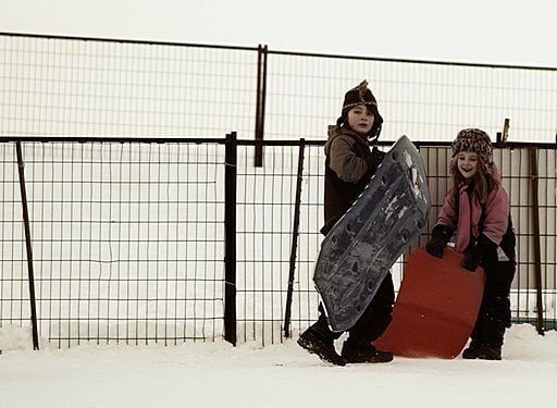 two kids standing in the snow with their sleds