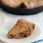 A slice of chocolate chip skillet cookie cake on a white plate, with the rest of the cookie cake in a black pan in the background. The cake appears dense and filled with gooey chocolate chips.