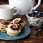 Three cinnamon rolls on a blue plate sit next to a bowl of blueberries, a white mug, and a white teapot on a rustic wooden table—with hints of Philly chocolate cream cheese swirled inside each roll. Some loose blueberries are scattered near the bowl.
