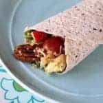 A close-up of a quinoa wrap filled with scrambled eggs, grape tomatoes, spinach, a pecan, and shredded cheese, served on a light blue plate with a floral tablecloth beneath.