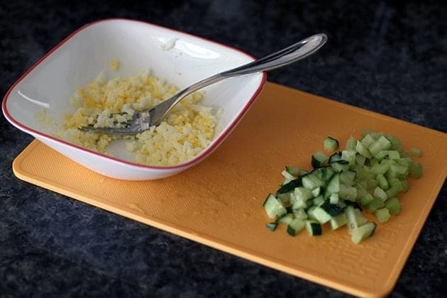 smashing the hard boiled egg in a bowl using a fork, chopped cucumber on the side