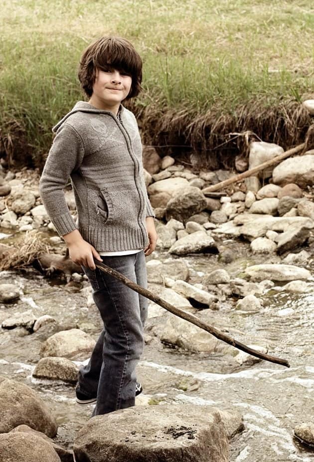 young boy holding a wood stick while standing in a stone on the creek