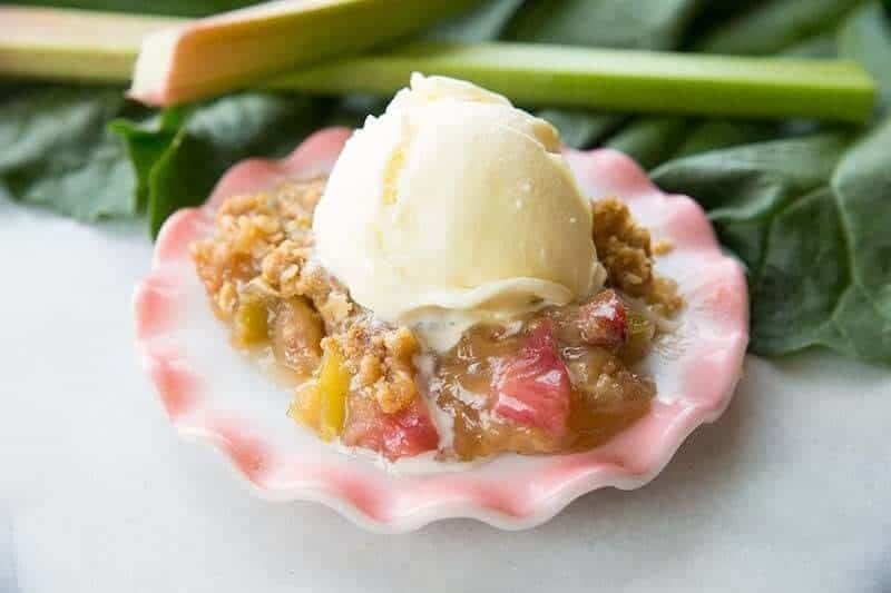 A serving of rhubarb crisp topped with a scoop of vanilla ice cream sits on a pink-rimmed plate, reminiscent of classic Strawberry Recipes, with fresh rhubarb stalks and green leaves in the background.