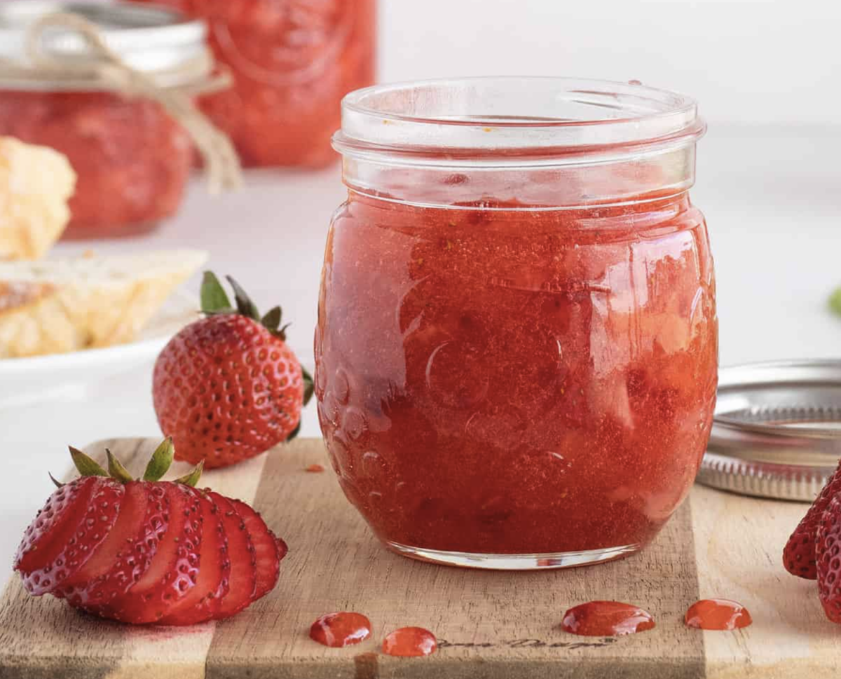 A jar of strawberry jam sits on a wooden board, surrounded by fresh and sliced strawberries, with a metal lid nearby. In the background, there are more jars and a blurred plate of food—an inviting scene perfect for anyone gathering inspiration for their next strawberry recipes.