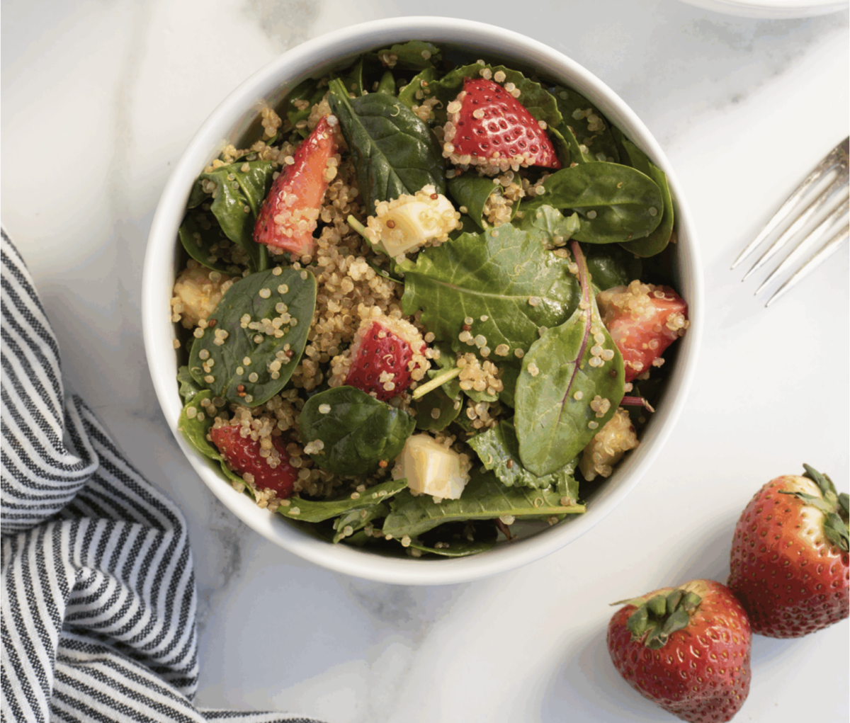 A bowl of quinoa salad with spinach, chopped strawberries, and cheese sits on a white surface next to a striped napkin, a fork, and two whole strawberries, making it a fresh addition to your favorite Strawberry Recipes.