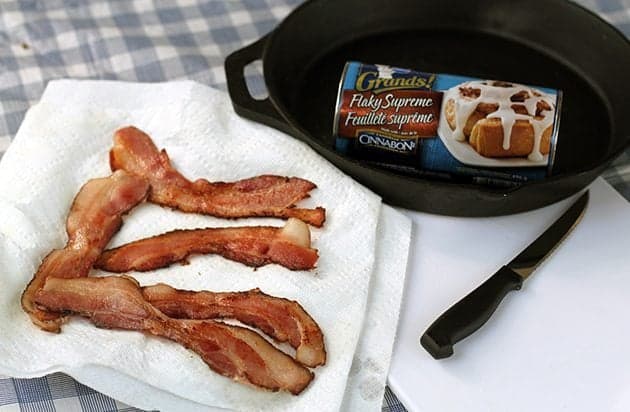 five slices of cooked bacon in a paper towel, a can of Pillsbury Grands! Cinnamon rollss in a skillet and a knife